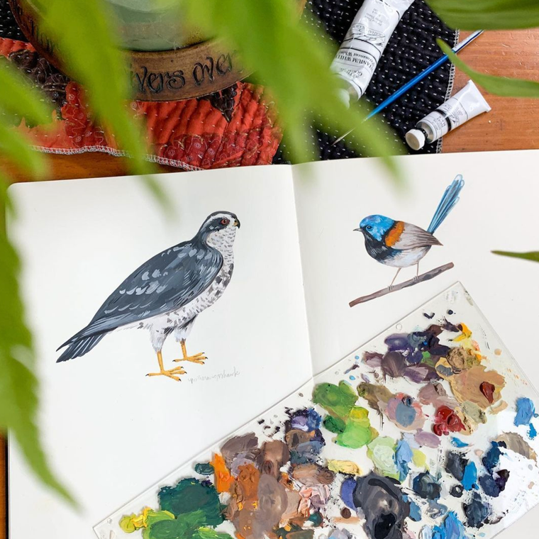 Open sketchbook on a table with a colorful paint palette, a painted hawk, and a painted fairy wren